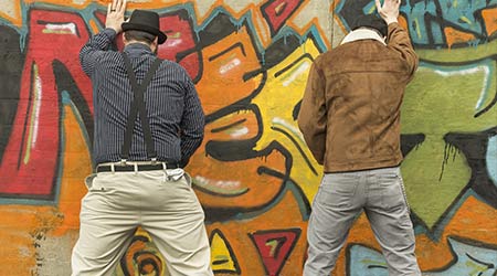 Two hipster men urinating in public on a wall featuring a painted portrait