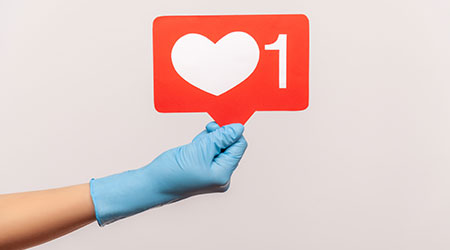 Profile side view closeup of human hand in blue surgical gloves holding social media like stick. indoor, studio shot, isolated on gray background.