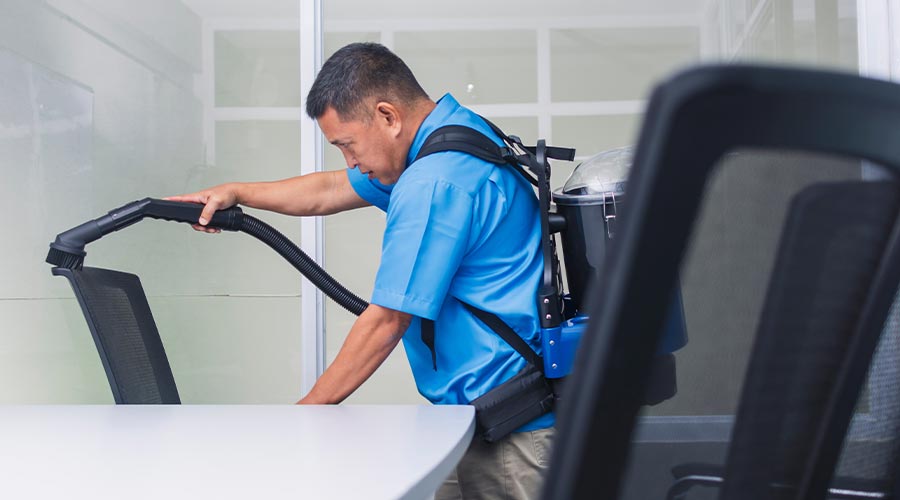 A professional janitor in blue uniform using a commercial backpack vacuum to clean a modern office chair in a bright workspace.