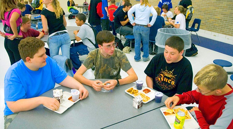 High school students gather and socialize together as they eat lunch in a high school cafeteria lunchroom.