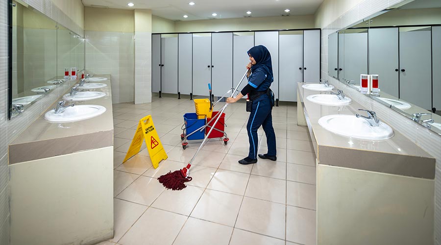 a hijab female employee in uniform is cleaning the interior of a public toilet in a commercial building