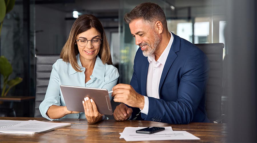 Team of diverse partners mature Latin business man and European business woman discussing project on tablet sitting at table in office.